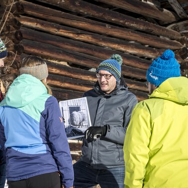 Eine Gruppe von fünf Personen steht im Schnee vor einer Holzscheune. Sie betrachten gemeinsam ein Foto, das einer der Teilnehmer in der Hand hält. | © Kleinwalsertal Tourismus | Dominik Berchtold