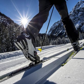 Ein Skifahrer gleitet über eine schneebedeckte Langlaufstrecke. Im Hintergrund leuchten die Berge unter einem klaren blauen Himmel.