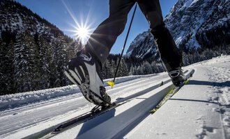 Ein Skifahrer gleitet über eine schneebedeckte Langlaufstrecke. Im Hintergrund leuchten die Berge unter einem klaren blauen Himmel.