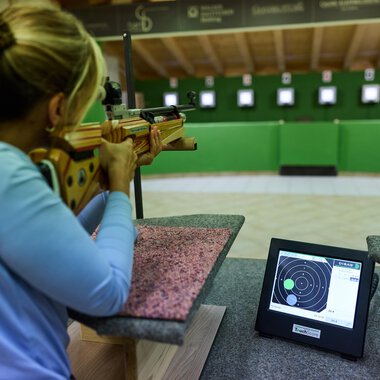 A marksman aims a rifle at a target. In the foreground, a monitor is visible displaying the results. | © Kleinwalsertal Tourismus | Oliver Farys