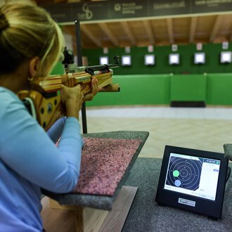 A marksman aims a rifle at a target. In the foreground, a monitor is visible displaying the results. | © Kleinwalsertal Tourismus | Oliver Farys