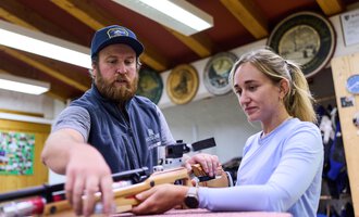 A man is showing a woman how to use an air rifle. Both appear focused in a sporty environment. | © Kleinwalsertal Tourismus | Oliver Farys