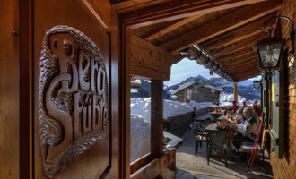 Ein gemütlicher Gasthof mit Holzverzierungen und Blick auf schneebedeckte Berge. Draußen sitzen Gäste an einem Tisch und genießen die alpine Atmosphäre. | © Bergstüble | Michaela Beck Dornach