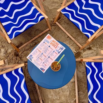 A group of blue loungers stands around a round table in the sand. On the table is a drink menu and a cocktail. | © MORE VIBES by Alex Lambsdorff