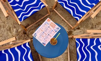 A group of blue loungers stands around a round table in the sand. On the table is a drink menu and a cocktail. | © MORE VIBES by Alex Lambsdorff