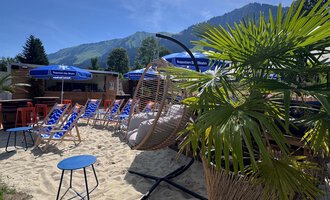 A relaxed beach area with sun loungers and a hammock.  
Surrounded by palm trees and mountains under a clear sky. | © MORE VIBES by Alex Lambsdorff