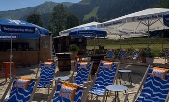A sunny beach with blue deck chairs and beach umbrellas. In the background, green mountains can be seen. | © MORE VIBES by Alex Lambsdorff