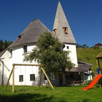 Ein charmantes, weißes Gebäude mit einem spitzen Dach, umgeben von Wiesen und Bäumen. Im Vordergrund stehen eine Schaukel und eine Rutsche, die zum Spielen einladen.