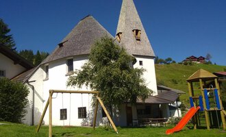 Ein charmantes, weißes Gebäude mit einem spitzen Dach, umgeben von Wiesen und Bäumen. Im Vordergrund stehen eine Schaukel und eine Rutsche, die zum Spielen einladen.