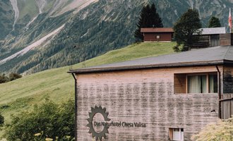 A modern building in the mountains with a wooden facade. The surroundings are green with an impressive mountain landscape in the background. | © Das Naturhotel Chesa Valisa