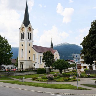 Eine malerische Kirche mit einem hohen Turm steht im Mittelpunkt eines kleinen Platzes. Im Hintergrund sind grüne Bäume und Berge zu sehen.