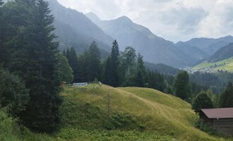 Eine malerische Landschaft mit grünen Hügeln und hohen Bergen. Im Vordergrund steht eine kleine Hütte umgeben von Bäumen. | © Nicole Lughammer