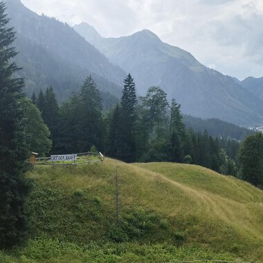 A picturesque landscape with green hills and tall mountains. In the foreground, there is a small cabin surrounded by trees. | © Nicole Lughammer