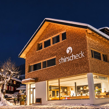 Ein modernes Gebäude mit einer Holzfassade und großen Fenstern, das bei Nacht beleuchtet ist. Es ist von Schnee umgeben und liegt in einer malerischen winterlichen Umgebung.