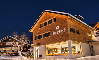 Ein modernes Gebäude mit einer Holzfassade und großen Fenstern, das bei Nacht beleuchtet ist. Es ist von Schnee umgeben und liegt in einer malerischen winterlichen Umgebung.
