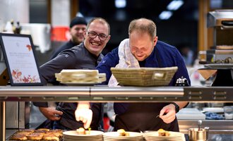 Two chefs are smiling in a kitchen as they prepare dishes. In the background, another chef can be seen, who also appears to be cheerful.