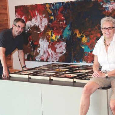 A man and a woman are standing at a table on which numerous picture frames are lying. In the background, colorful abstract paintings can be seen. | © Gerti Maitz