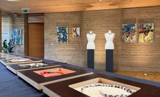 A modern exhibition with several pieces of jewelry in display cases. In the background, there are mannequins and colorful paintings on the wall. | © Gerti Maitz