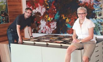 A man and a woman are standing at a table on which numerous picture frames are lying. In the background, colorful abstract paintings can be seen. | © Gerti Maitz