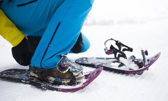 Eine Person schnallt Schneeschuhe an, um im Schnee zu wandern. Die Umgebung ist winterlich und schneebedeckt. | © Kleinwalsertal Tourismus | Oliver Farys