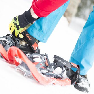 Eine Person schnallt sich Schneeschuhe an, während sie im Schnee steht. Der Boden ist weiß und die Kleidung ist bunt. | © Kleinwalsertal Tourismus eGen | Oliver Farys