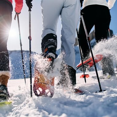 Drei Personen wandern im Schnee mit Schneeschuhen. Die Sonne scheint und Schnee spritzt von ihren Bewegungen auf. | © Kleinwalsertal Tourismus | Oliver Farys