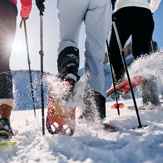 Drei Personen wandern im Schnee mit Schneeschuhen. Die Sonne scheint und Schnee spritzt von ihren Bewegungen auf. | © Kleinwalsertal Tourismus | Oliver Farys