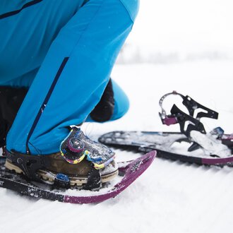 A person is putting on snowshoes while sitting on a snow-covered ground. The equipment and clothing are suitable for winter outdoor activities. | © Kleinwalsertal Tourismus | Oliver Farys