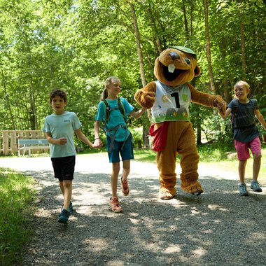 Children are happily walking along a path through the forest, accompanied by a cheerful mascot. The atmosphere is friendly and inviting. | © Kleinwalsertal Tourismus | Oliver Farys
