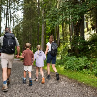 A family is hiking on a forest path. Surrounded by tall trees and green vegetation, they enjoy nature. | © Kleinwalsertal Tourismus | Oliver Farys