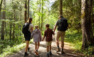 A family is hiking together on a forest path. They are enjoying the nature around them among tall trees. | © Kleinwalsertal Tourismus | Oliver Farys