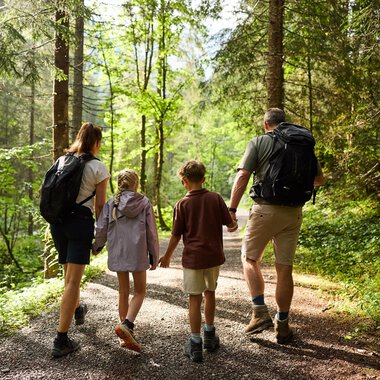 A family is hiking together on a forest path. They are enjoying the nature around them among tall trees. | © Kleinwalsertal Tourismus | Oliver Farys
