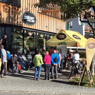 A group of people stands in front of a café with a yellow awning. It is a sunny day, and some drivers are waiting while other cyclists are parking their bikes. | © Schönegger Käsealm
