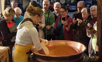A person is preparing something in a large pot under a tent. In the background, a group of people is watching with interest. | © Schönegger Käsealm