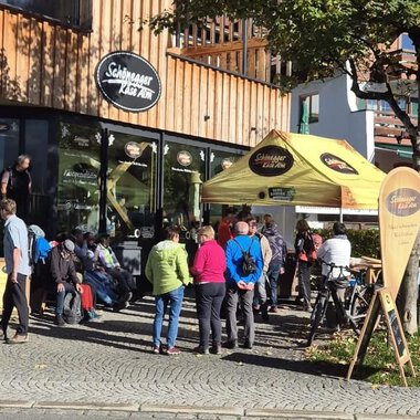 A group of people stands in front of a café with a yellow awning. It is a sunny day, and some drivers are waiting while other cyclists are parking their bikes. | © Schönegger Käsealm