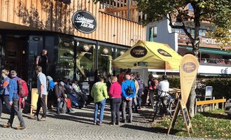 A group of people stands in front of a café with a yellow awning. It is a sunny day, and some drivers are waiting while other cyclists are parking their bikes. | © Schönegger Käsealm