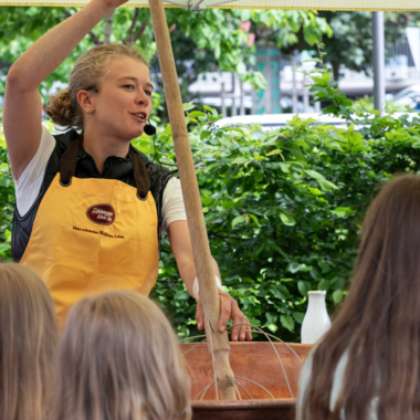 A woman is explaining something with a long stick in her hand. In the background, children can be seen listening intently. | © Schönegger Käsealm Susanne Lang