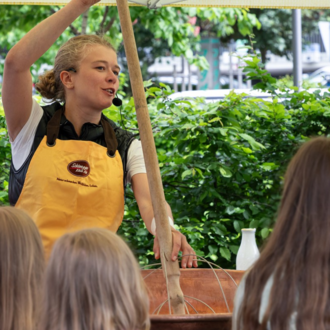 A woman is explaining something with a long stick in her hand. In the background, children can be seen listening intently. | © Schönegger Käsealm Susanne Lang
