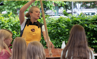 A woman is explaining something with a long stick in her hand. In the background, children can be seen listening intently. | © Schönegger Käsealm Susanne Lang