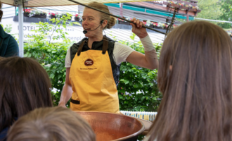 A woman in a yellow apron is explaining something in front of a group of listeners. In the background, a green hedge and a covered market area can be seen. | © Schönegger Käsealm Susanne Lang