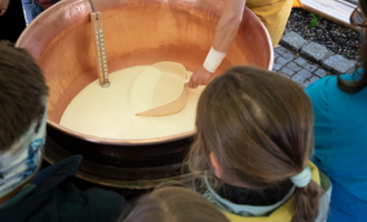 A person is stirring a mixture in a large copper kettle. In the background, there are interested spectators, including children, who are watching curiously. | © Schönegger Käsealm Susanne Lang