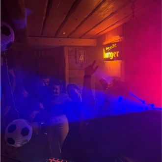 Eine lebhafte Party in einem gemütlichen Raum mit bunten Lichtern und Nebel. Menschen tanzen und scheinen viel Spaß zu haben.