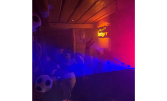 Eine lebhafte Party in einem gemütlichen Raum mit bunten Lichtern und Nebel. Menschen tanzen und scheinen viel Spaß zu haben.
