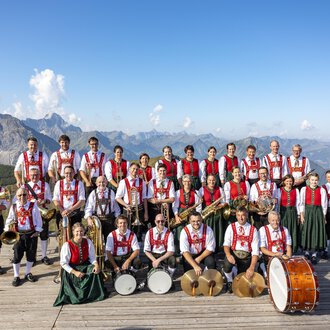 Eine Musikkapelle in traditioneller Tracht steht auf einer Plattform in den Bergen. Im Hintergrund sind beeindruckende Berge und blauer Himmel zu sehen. | © Trachtenkapelle Riezlern