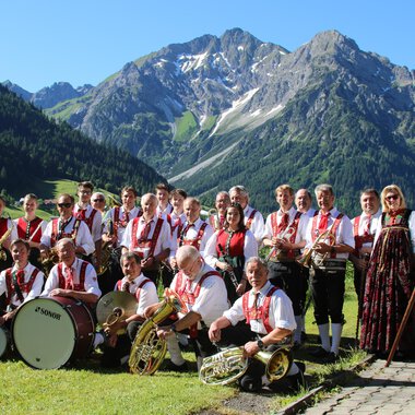A music band in traditional attire stands in front of a picturesque mountainous landscape. In the background, green meadows and snow-capped mountains can be seen. | © Musikverein d´Hirschegger | Jockl Metzger