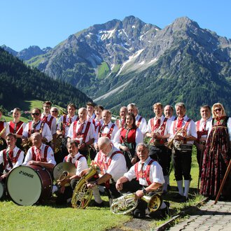 A music band in traditional attire stands in front of a picturesque mountainous landscape. In the background, green meadows and snow-capped mountains can be seen. | © Musikverein d´Hirschegger | Jockl Metzger