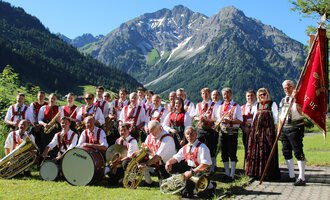 A music band in traditional attire stands in front of a picturesque mountainous landscape. In the background, green meadows and snow-capped mountains can be seen. | © Musikverein d´Hirschegger | Jockl Metzger