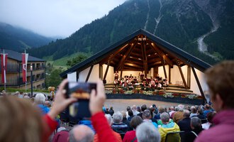 An outdoor stage in the mountains with an orchestra performance. Many spectators sit in front and enjoy the music in the twilight. | © Kleinwalsertal Tourismus | Oliver Farys