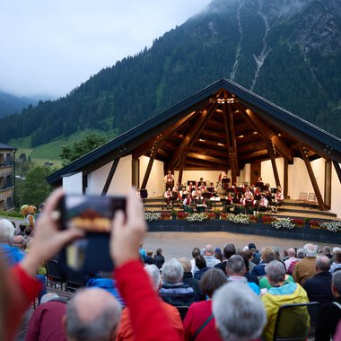 An outdoor stage in the mountains with an orchestra performance. Many spectators sit in front and enjoy the music in the twilight. | © Kleinwalsertal Tourismus | Oliver Farys