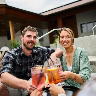 A cheerful couple enjoying cocktails together. Cozy seating is visible in the background. | © Kleinwalsertal Tourismus | Oliver Farys